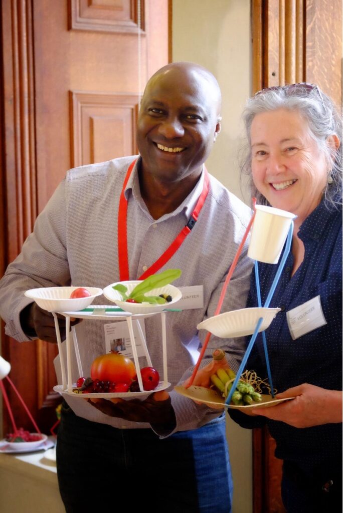 Two smiling volunteers, a black man wearing a lanyard and a white woman with long white hair, hold two fruit stands made on the Fitzwilliam Family Day.