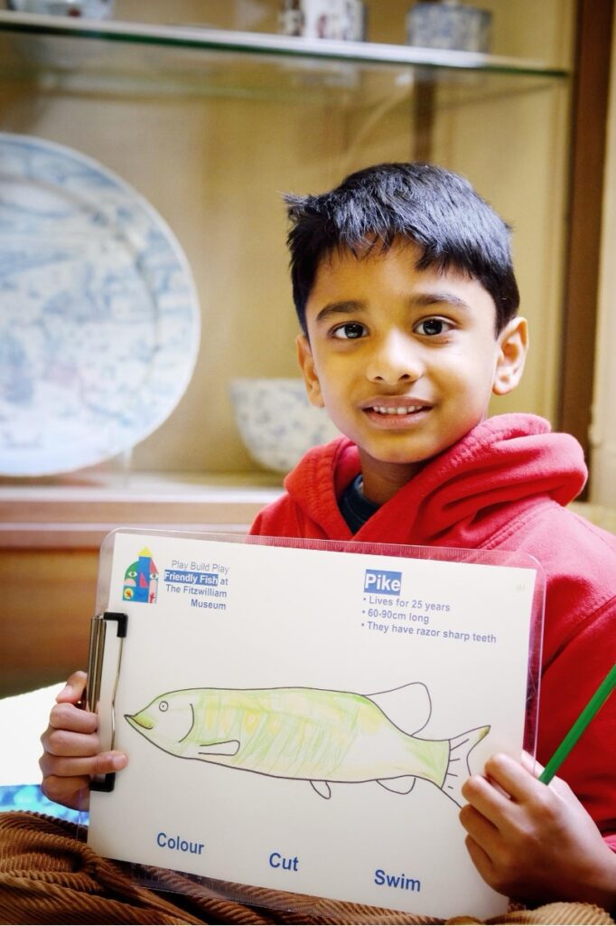 A young boy in a red hoodie holds up his drawing of a fish in a gallery at the Fitzwilliam Museum.