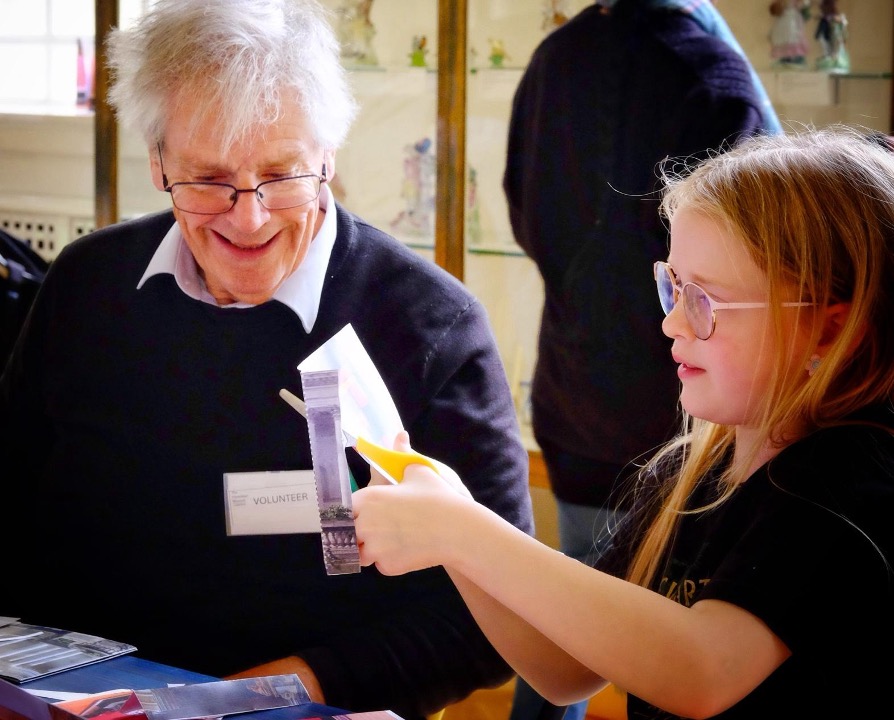 An older man wearing glasses and a Volunteer badge sits alongside a young girl at a table in the Museum. The girl is cutting a piece of paper with scissors.