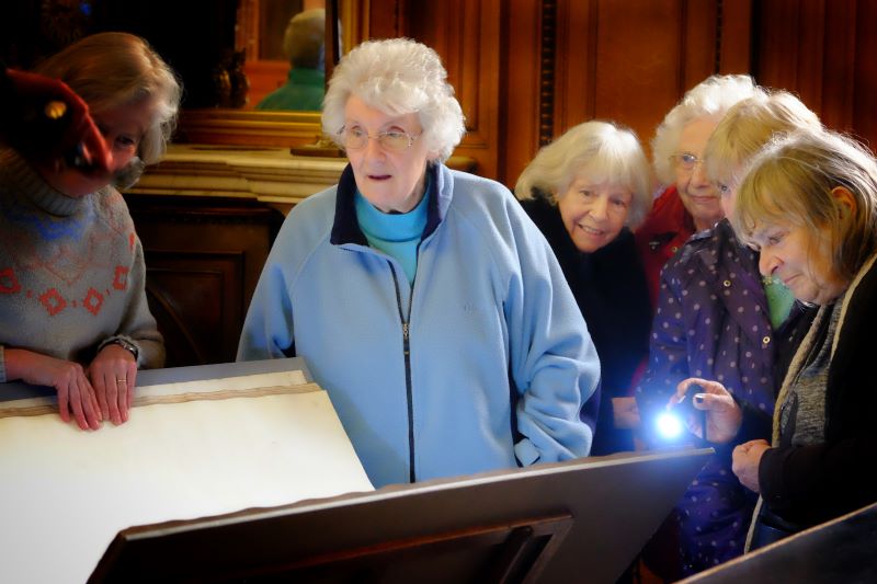 Group of older women stand together looking at a large open book.