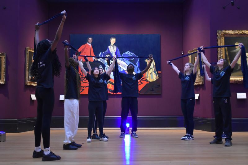 Group of young people stand in a semi-circle in a gallery holding a band above their heads