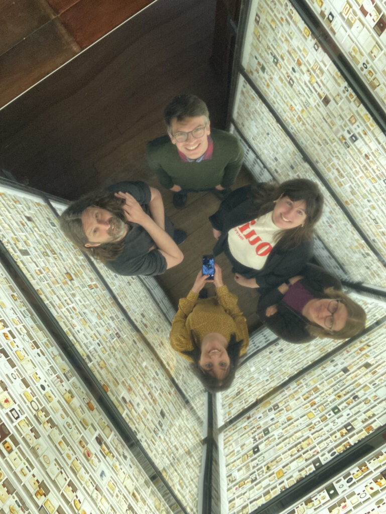 Five adults standing in the Grant Museum's Micrarium (room of slides) taking a selfie looking up at the reflective ceiling.