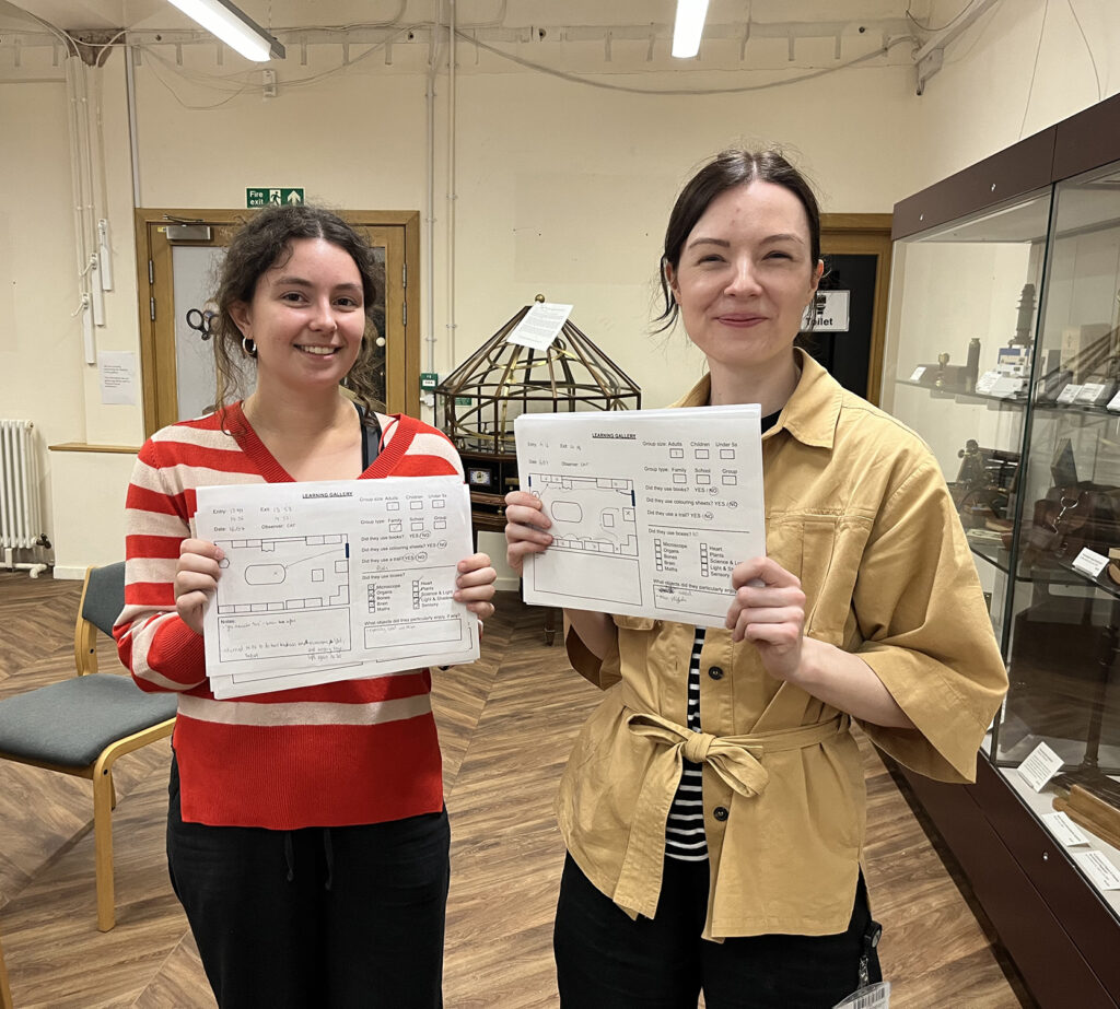 Two women hold smile and hold up tracking sheets in the Whipple Museum learning gallery.