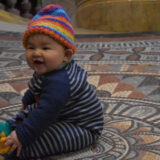 A baby sitting on the mosaic floor of the Museum wearing a mosaic hat.