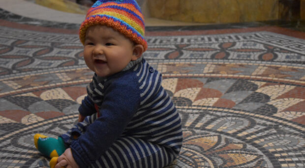 A baby sitting on the mosaic floor of the Museum wearing a mosaic hat.