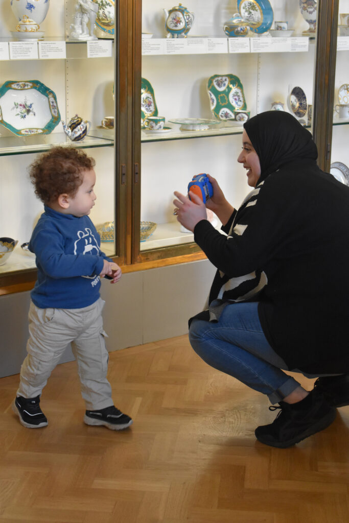 An adult crouches and uses a children’s camera to take a picture of a child in front of display cases at the museum.