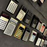 A collection of calculators lined across a table with a black tablecloth.