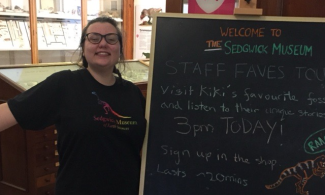 A member of staff smiling next to their tour advert, on a museum chalk board