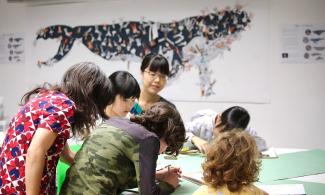 Several children and adults stand with their back to the camera, gathered around a table where they are drawing and making with clay