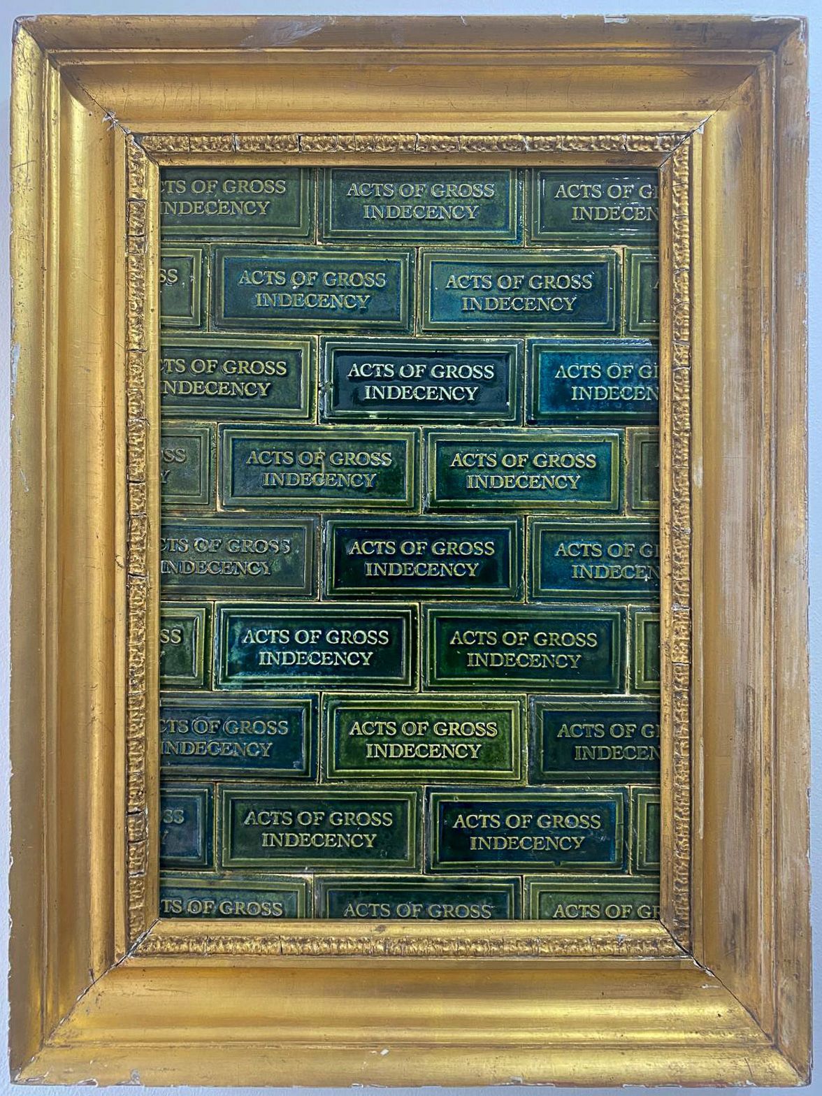 A gold frame filled with green earthenware tiles which say 'Acts of gross indecency'