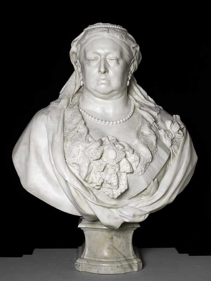 White marble bust of Queen Victoria, wearing a veil and an elaborate frilly dress, with a string of pearls. She look solemn and is shown in late middle age with wrinkles and a jowlly chin. The marble is very smooth, bright white, and shiny.
