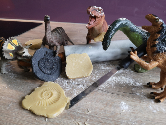 Five plastic dinosaurs with a marble rolling pin. Two on the left are holding a black plastic ammonite fossil that they have just pressed into a piece of cookie dough