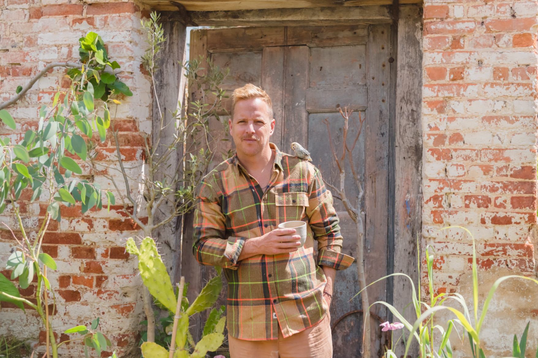 The artist Mark Mann standing outside with a bird perched on his shoulder.