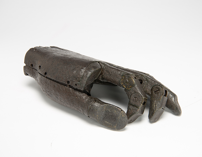 iron prosthetic hand side view. It is a left hand, and lies palm down with the thumb underneath, with the fingers slightly curled to demonstrate their articulation. The hand ends at the wrist and is made of dark iron and the four fingers are fully articulated, while the thumb is not jointed.