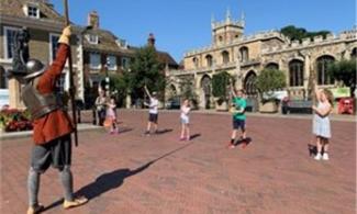 Adult dressed as an English Civil War soldier in large outdoor square instructs 5 children holding batons