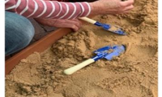 Photograph of 2 people crouching next to a sandpit containing trowels. Lower half of bodies only visible.