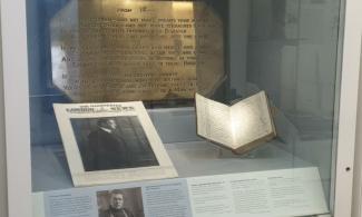 Photograph of a display case featuring a brass plaque (background), a paper document wtih a photograph and an open paper diary.