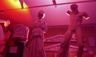 Two statues looking towards one another backlit with red. The left is a woman, wearing a robe and helmet, the right is a naked satyr dancing.