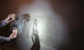 A photograph showing children's hands holding up a piece of perspex. They are using a torch to create a shadow with the perspex, which they have drawn shapes on.