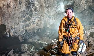  A portrait photograph of woman Earth Scientist wearing caving clothing and equipment, surrounded by rocks in a cave.