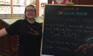A member of museum staff enthusiastically smiling next to a chalk board sign advertising her tour.