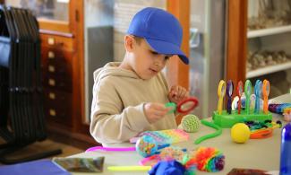 Child in the center of the image wearing a beige hoodie and bright blue cap they are holding a magnifying glass. There is a table in front with a variety of colorful sensory and fidget toys on. In the background there are several cabinets containing items from the Sedgwick Museum. 