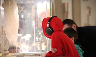 A child in a red hoodie, wearing ear defenders looks into a glass cabinet containing specimens from the Zoology Museum. To the left is the bottom half of a taxidermic penguin and behind the child is another child and an adult.  