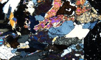 A 'thin section' of the mineral eclogite, as viewed down a petrographic microscope. It is a slice of rock, with areas of varied colours (different minerals), mainly black, grey, blue, pink, orange with small amounts of purple and yellow.