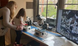  A mum helps a young girl look down a microscope, standing on a stool. The table has a second microscope and a large screen, showing white, shell-like fragments which are tiny fossils, the size of a grain of sand.
