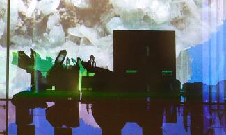 A projection of a white crystal mineral on a blue background shines through a museum display case of minerals. The minerals and a square display labels are silhouetted in black against the bright image.