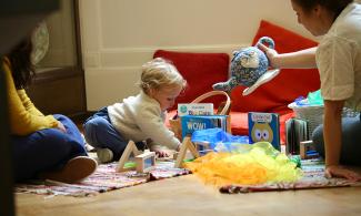A young child sits on a rug in a Museum gallery and plays with toys. Two adults sit either side of the child.