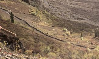 A young geologist walks in the foreground of a rocky terrain, smiling with a pick-axe over her shoulder.