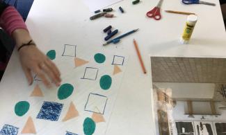 Children leaning over a table, which is covered in pencils, scissors, oil pastel and pieces of artwork. The artwork is the bottom left corner has a light green background, with peach triangles, dark green circles and navy blue squares drawn on top of it.