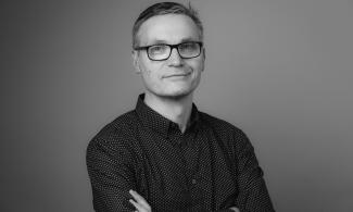 A black and white photograph of a man with short hair and desk glasses looking at the camera.