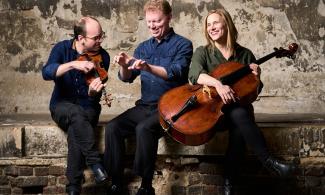 Three people sat in a row, the man on the left holding a violin, and the woman on the right holds a cello. 