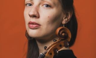 A woman with long brown hair looking directly at the camera, holding a violin. The background is orange.