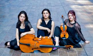 Three woman sit on a paved floor, with a cello in front of them.