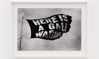 A black and white photograph of a flag rustling in the wind. The flag reads 'Here is a gale warning'