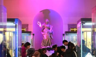 A museum room with a background of soft purple light, where families with children enjoy an activity on a blue table, sitting on yellow chairs.