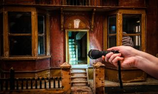 A hand holds a torch which light shines towards a miniature house.