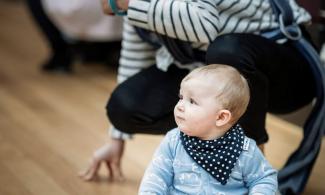 alt='a baby is sat on the floor in a blue shirt, his mother crouches behind"