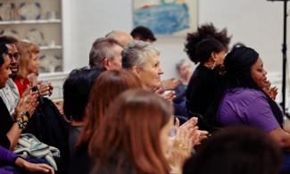 A group of people sat in rows applauding an event in the Kettle's Yard house. The image is focused on a white woman with short grey hair.