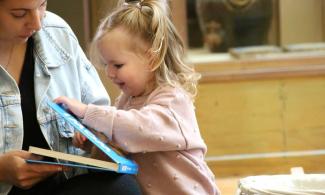 alt= "a young girl in a pink jumper opens a story book with her mum on her right"