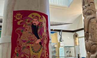 A pillar in the museum gallery featuring a predominantly red and gold poster celebrating the Lunar New Year. It depicts a bearded figure holding a bladed weapon and Chinese characters. In the background are display cases and to the right of the image can be seen a totem pole.