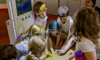 A group of children smile and reach to feel an object at the Science Centre, lit up with different coloured lights.