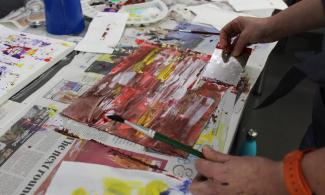 A busy desk covered with newspaper. On the top, a child holds an artworks of red, yellow and orange paint.
