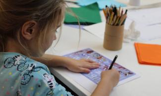 A young girl in a light blue t-shirt sit at a desk drawing.