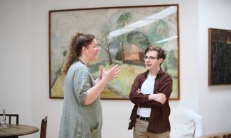 Two people speaking in the Kettle's Yard, a white woman with curly auburn hair wearing a sage green dress and a man with glasses and short brown hair, wearing a maroon shirt and cream trousers. 