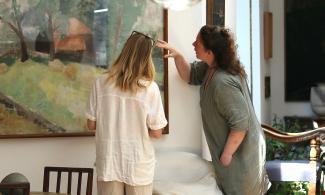 Two women in the Kettle's Yard house looking at a landscape artwork. One has blonde hair and wears a short-sleeved white shirt and cream linen trousers. The other person has curly auburn hair and wears a sage green dress.