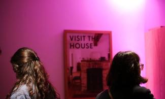 Two people stood in front of a poster of the Kettle's Yard house, illuminated by a pink light.